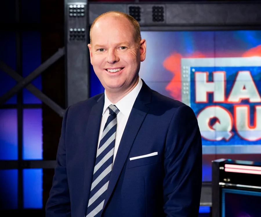 A man in a suit stands smiling in front of a colorful TV show set background.