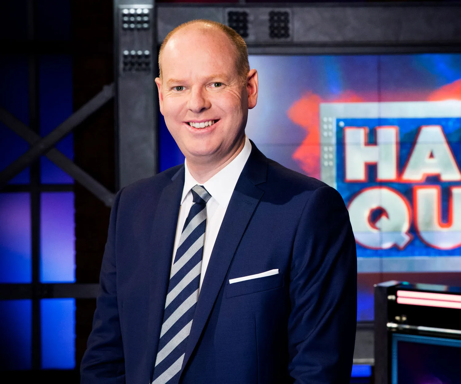 A man in a suit stands smiling in front of a colorful TV show set background.