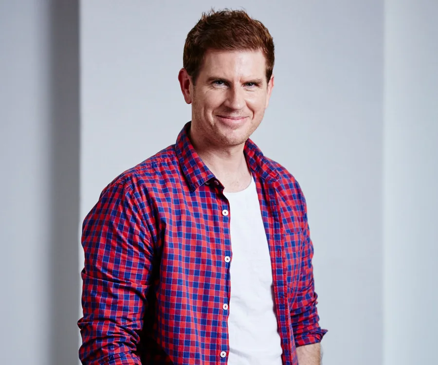 A man in a red and blue checkered shirt smiles at the camera in a neutral setting.
