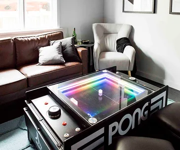 Retro Pong arcade table in a modern living room with a leather couch, armchair, and colorful LED lights.
