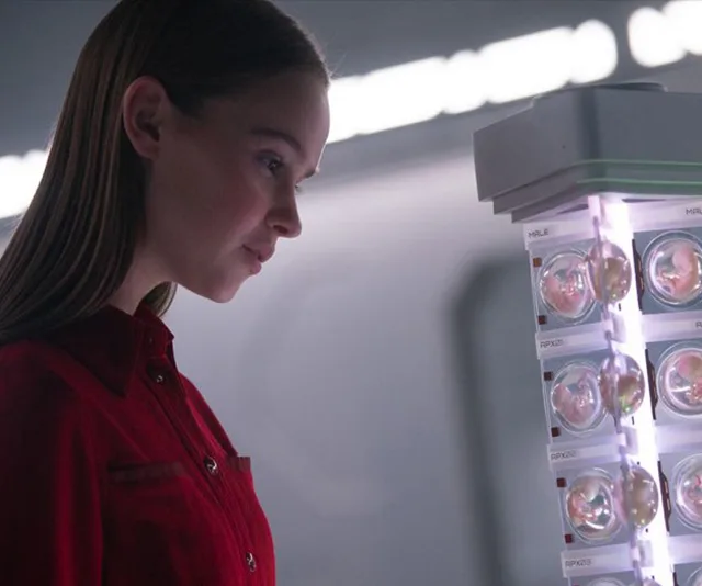 A young girl in a red shirt examines a futuristic device with individual compartments in a dimly lit room.