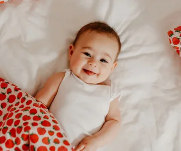 A smiling baby lies on a bed with white sheets and a strawberry-patterned blanket.