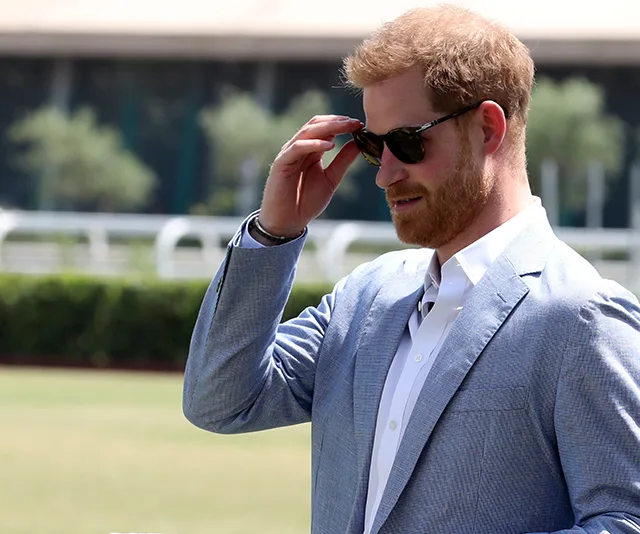 A man in a light blue suit and sunglasses stands outdoors, adjusting his glasses on a sunny day.
