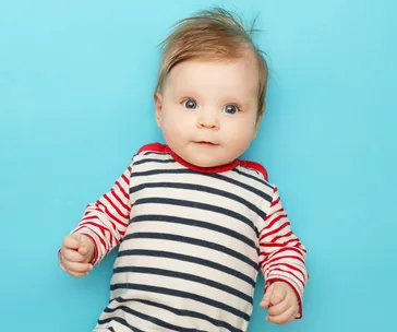 A baby in a striped shirt with red sleeves against a blue background, looking up with a curious expression.