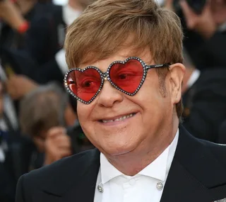 A person wearing heart-shaped, red-tinted sunglasses and a suit smiles at an event.
