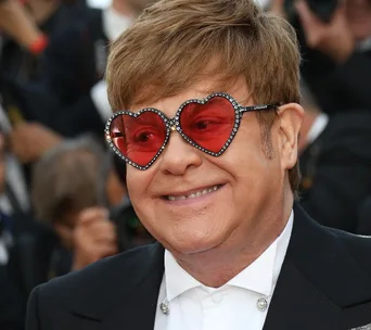 A person wearing heart-shaped, red-tinted sunglasses and a suit smiles at an event.
