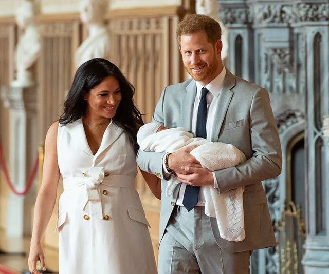 A couple smiling while holding a baby, with an ornate interior background.