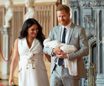 A couple smiling while holding a baby, with an ornate interior background.