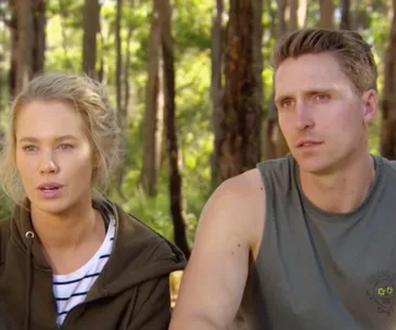 Two individuals sit outdoors in a forest setting, discussing seriously.