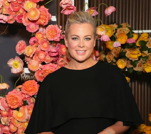 Samantha Armytage smiling in black dress, standing before colorful flower arrangement.