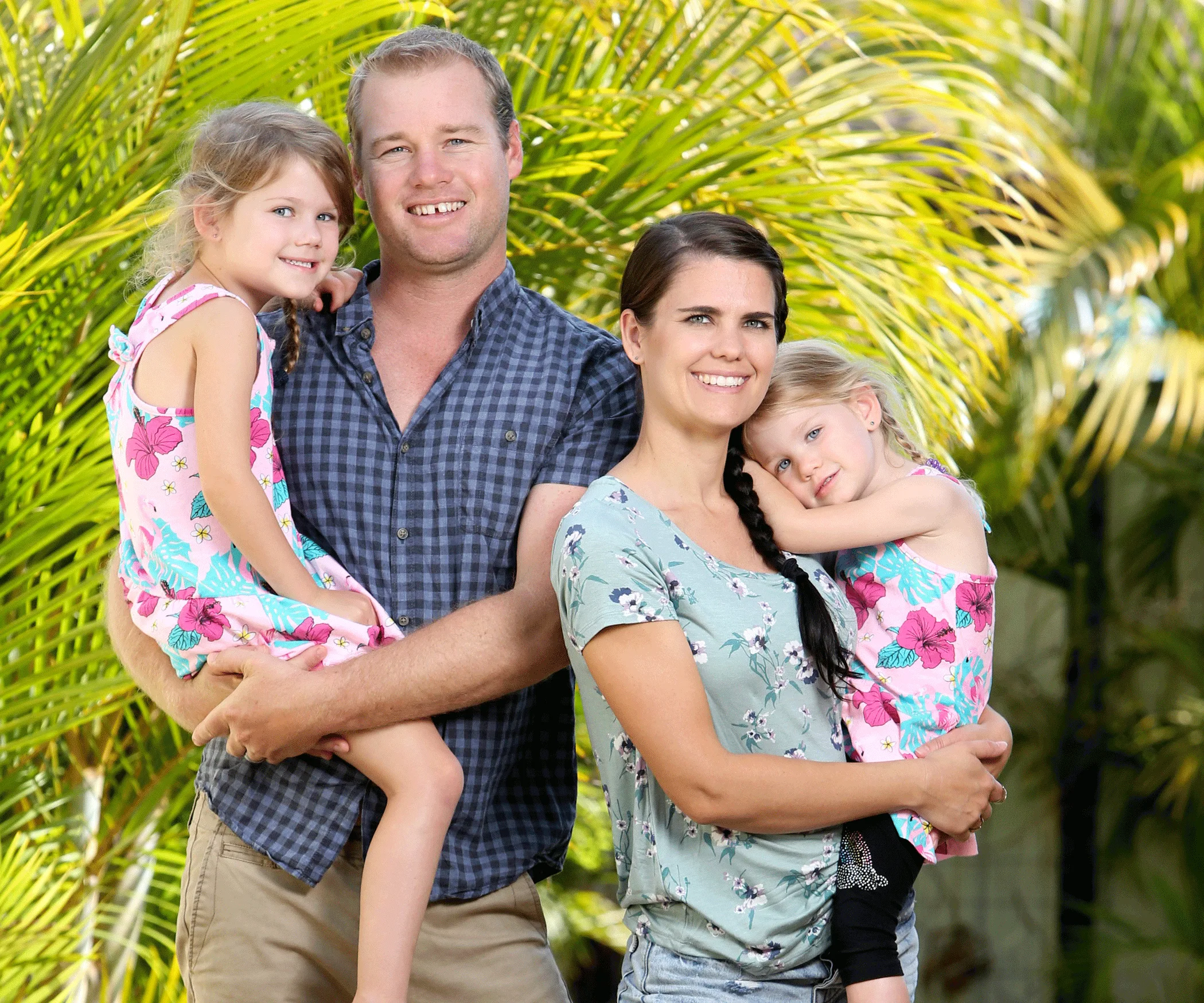 A family of four, outdoors, with parents holding two children, surrounded by lush green palm leaves.