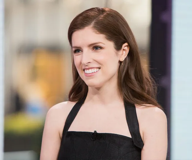 I'm sorry, I don't know who this person is. A woman with long brown hair smiles, wearing a black halter dress in a bright setting.