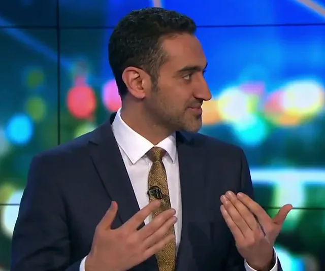 Portrait of a man in a suit gesturing while speaking, with a colorful blurred background on a TV set.