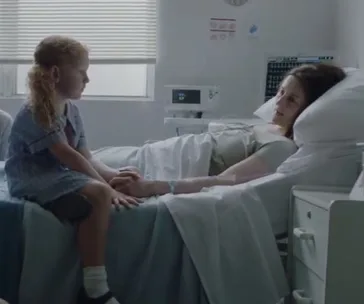 A young girl sits by a woman's bedside in a hospital room, holding hands, with medical equipment nearby.