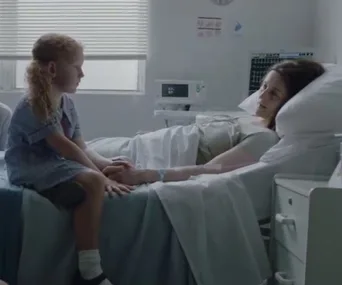 A young girl sits by a woman's bedside in a hospital room, holding hands, with medical equipment nearby.