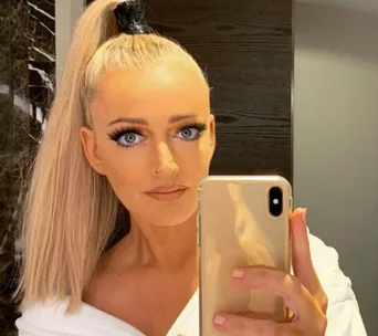 A person with long blonde hair in a high ponytail takes a selfie in a mirror using a smartphone.