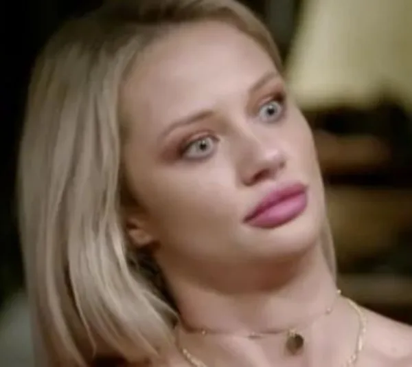 A surprised woman with blonde hair and wide eyes, wearing a necklace, looks sideways with a neutral background.