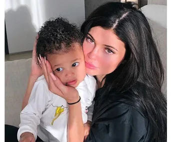 A woman with long dark hair lovingly holds a small child with curly hair, both seated indoors.