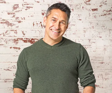 A person smiling in a green sweater against a white brick wall background.