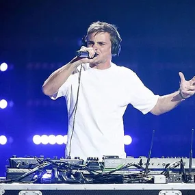 DJ performing live on stage, wearing headphones and holding a microphone, surrounded by equipment.