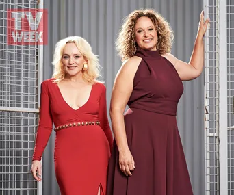 Two women from "Wentworth" Season 7 pose in elegant dresses against a metal fence backdrop.