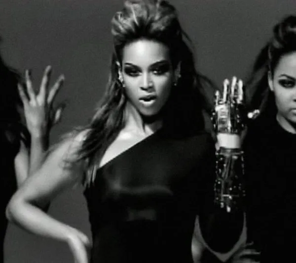 A woman in a black outfit performing a dance with backup dancers, raising her hand with emphasis.