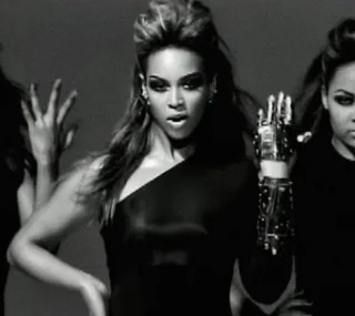 A woman in a black outfit performing a dance with backup dancers, raising her hand with emphasis.