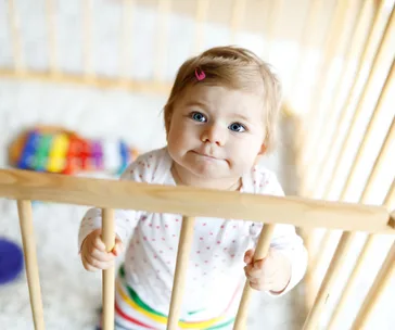 A baby with a pink hair clip stands in a wooden playpen, looking upward with toys visible in the background.