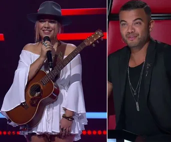 A singer with a guitar and a microphone on stage, and a judge watching attentively from a red chair.