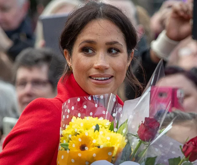 Woman in red coat holding a bouquet of flowers, surrounded by a crowd.