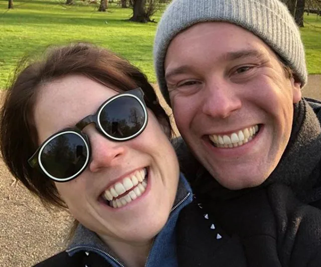 Two people smiling broadly for a sunny selfie in a park, one wearing sunglasses and the other a beanie.