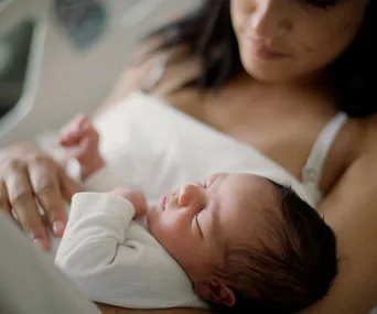 Mother holding a sleeping newborn baby on her chest, both are in a relaxed, comforting hospital setting.