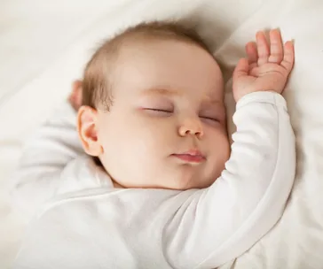 Baby sleeping peacefully on a white bed in a white onesie, arm raised above head.