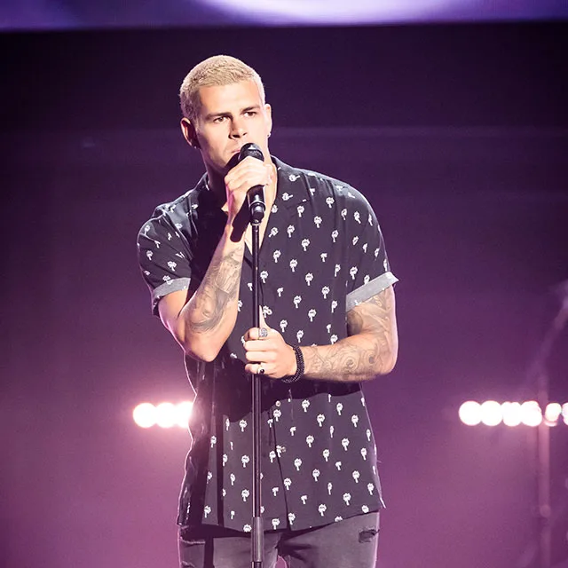 Singer performing on stage with short blonde hair and tattoos, wearing a patterned shirt, holding a microphone.