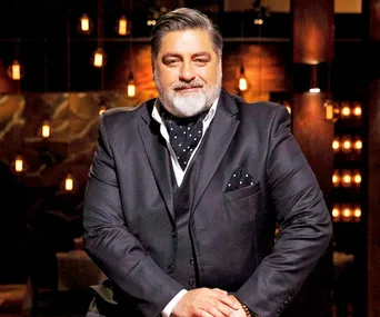 Matt Preston in a dark suit with a polka dot scarf and pocket square on a stylishly lit set with warm lights.