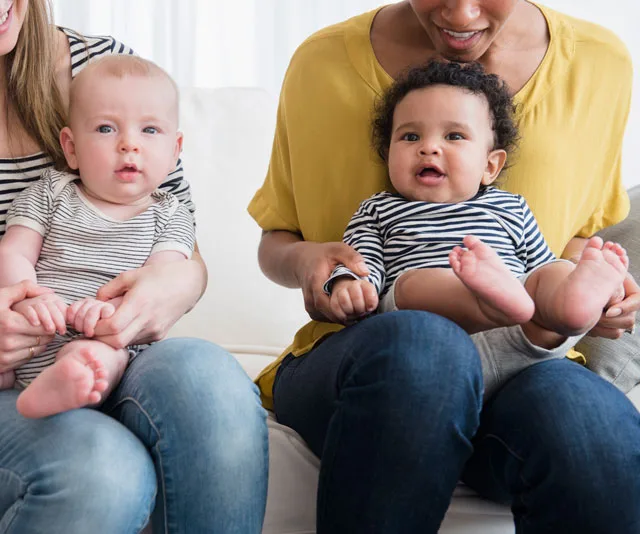 Two mothers sitting with their babies on their laps, both infants wearing striped shirts and smiling.