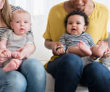 Two mothers sitting with their babies on their laps, both infants wearing striped shirts and smiling.