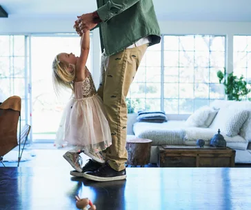 Toddler in a dress holding hands with an adult indoors in a sunlit living room.