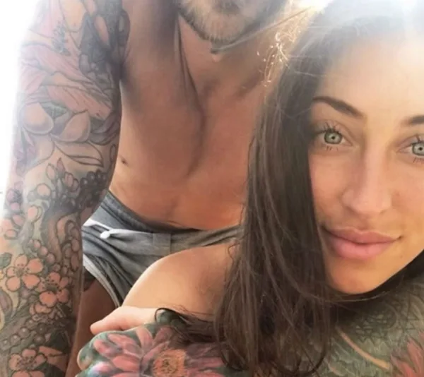A tattooed man and woman take a close-up selfie, smiling outdoors in sunlight.