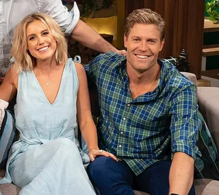 A smiling couple sits together on a couch, both casually dressed in light outfits.