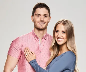 A couple smiling, the woman with long blonde hair wearing a blue top, and a man in a pink shirt with short brown hair.