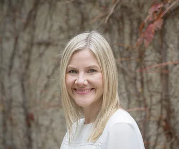 A smiling woman with blonde hair in front of a vine-covered wall.