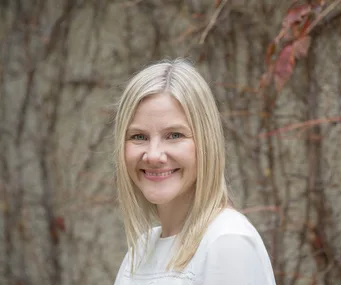 A smiling woman with blonde hair in front of a vine-covered wall.