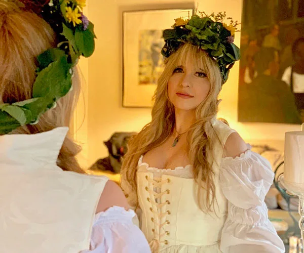 A woman in a white dress and floral crown gazes into a mirror, amid warm, artistic surroundings.
