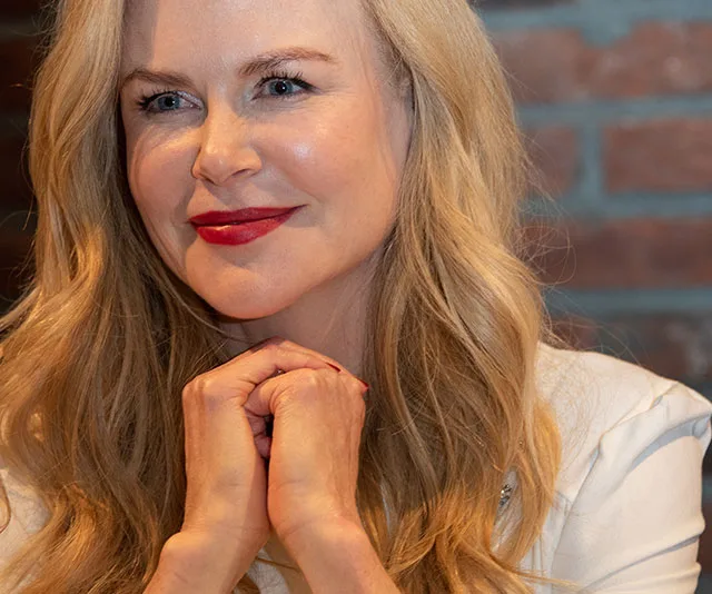 Nicole Kidman smiling with long blonde hair and red lipstick, hands clasped, in front of a brick wall.