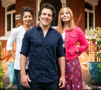 Three people smiling in front of a brick house, with plants around them.
