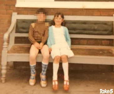 Two children sitting on a bench; one is pixelated, the other smiles, wearing a blue cardigan and white dress.