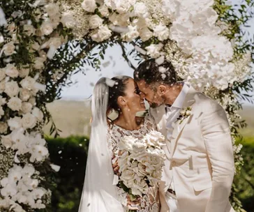 A couple kisses under a flower arch during their wedding ceremony, with petals falling around them.