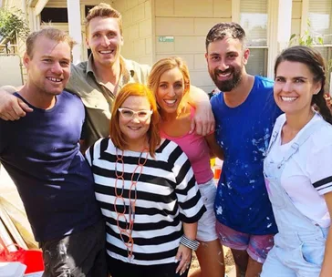 Group of six people smiling, outdoors, some in casual attire with paint splatters, in front of a house marked with "40".