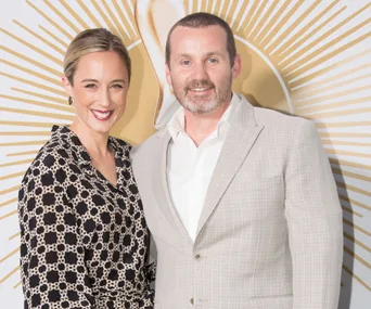 Two people posing together at an event, with a gold decorative background.